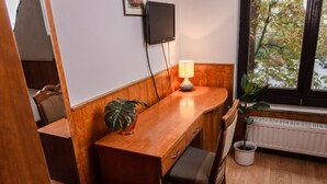 Una stanza compatta con una scrivania in legno, una sedia, un tavolino, una pianta in vaso, una lampada, un televisore fissato al muro e una finestra con vista sugli alberi.