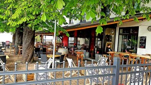Un caffè all'aperto con tavoli e sedie, un albero e un edificio con un cartello che riporta la scritta "HAPPY HOUR 16-18R".