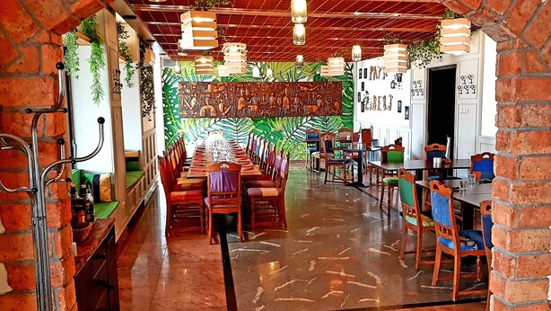 Un ristorante con un lungo tavolo comune, pareti in mattoni, piante sospese e un murale verdeggiante con motivi vegetali.