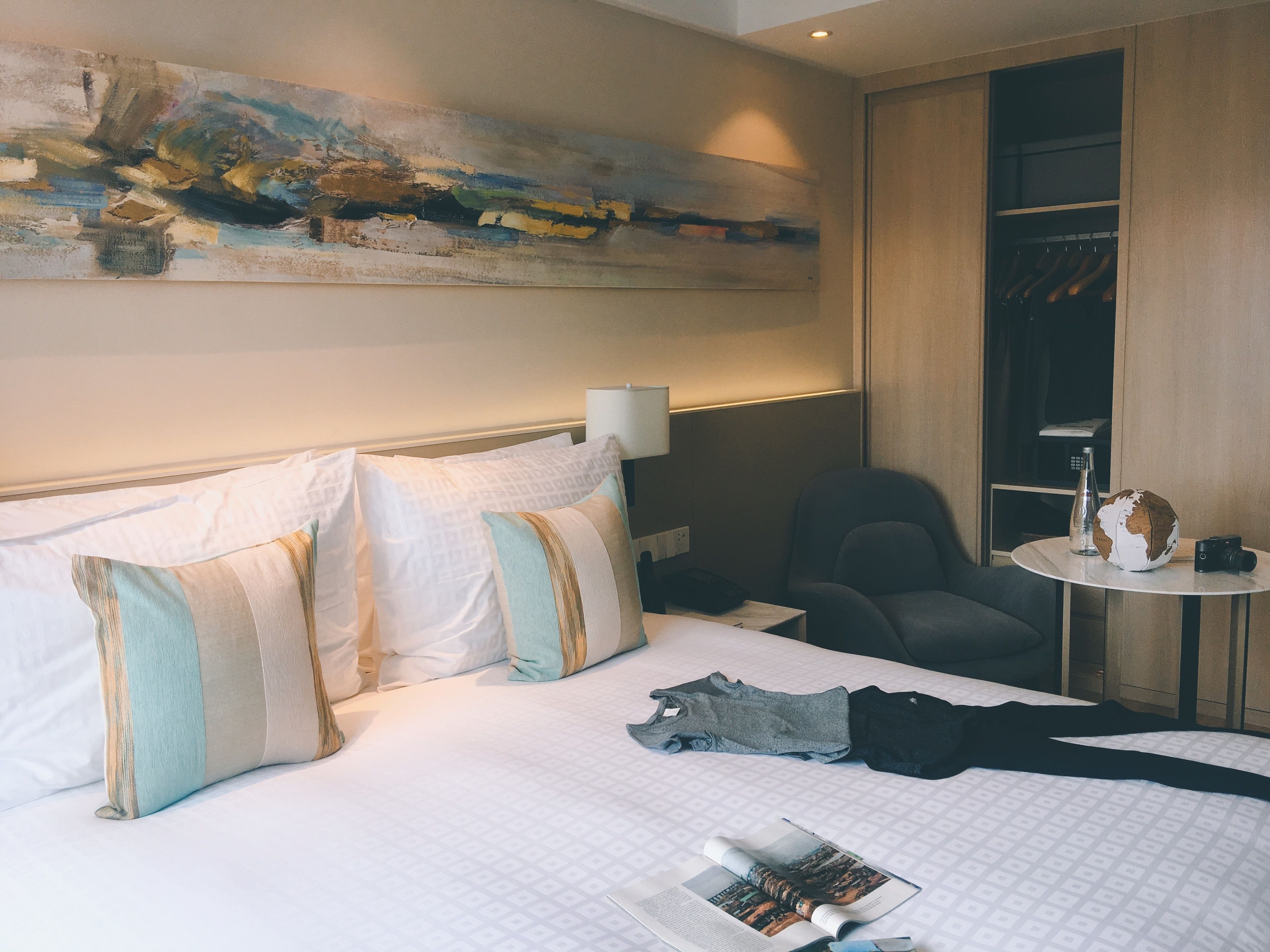 A hotel room with a neatly made bed, a chair, a small table with a globe and a camera, and a painting on the wall.