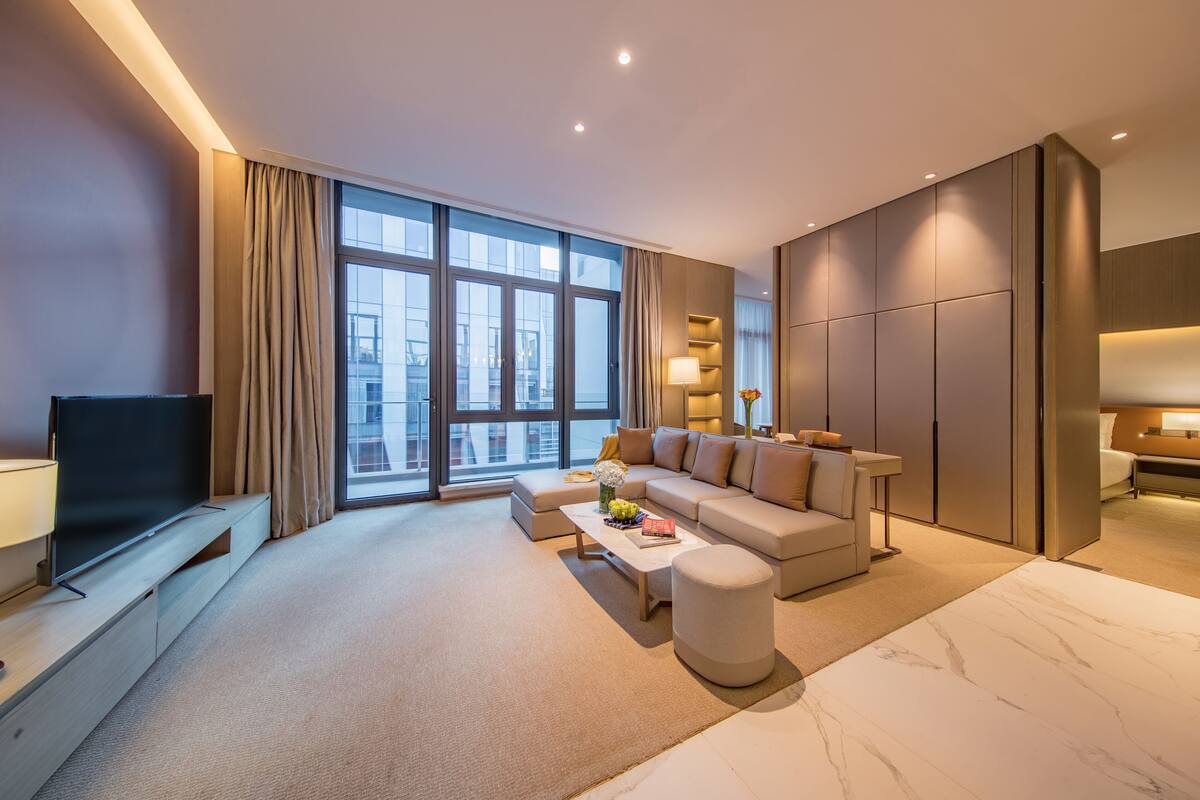 A modern hotel room with a large window, a sofa, a coffee table, and a TV.