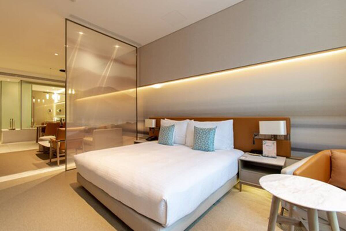 A modern hotel room with a large bed, a glass partition, a sitting area with a table, and a bathroom with a mirror and sink.