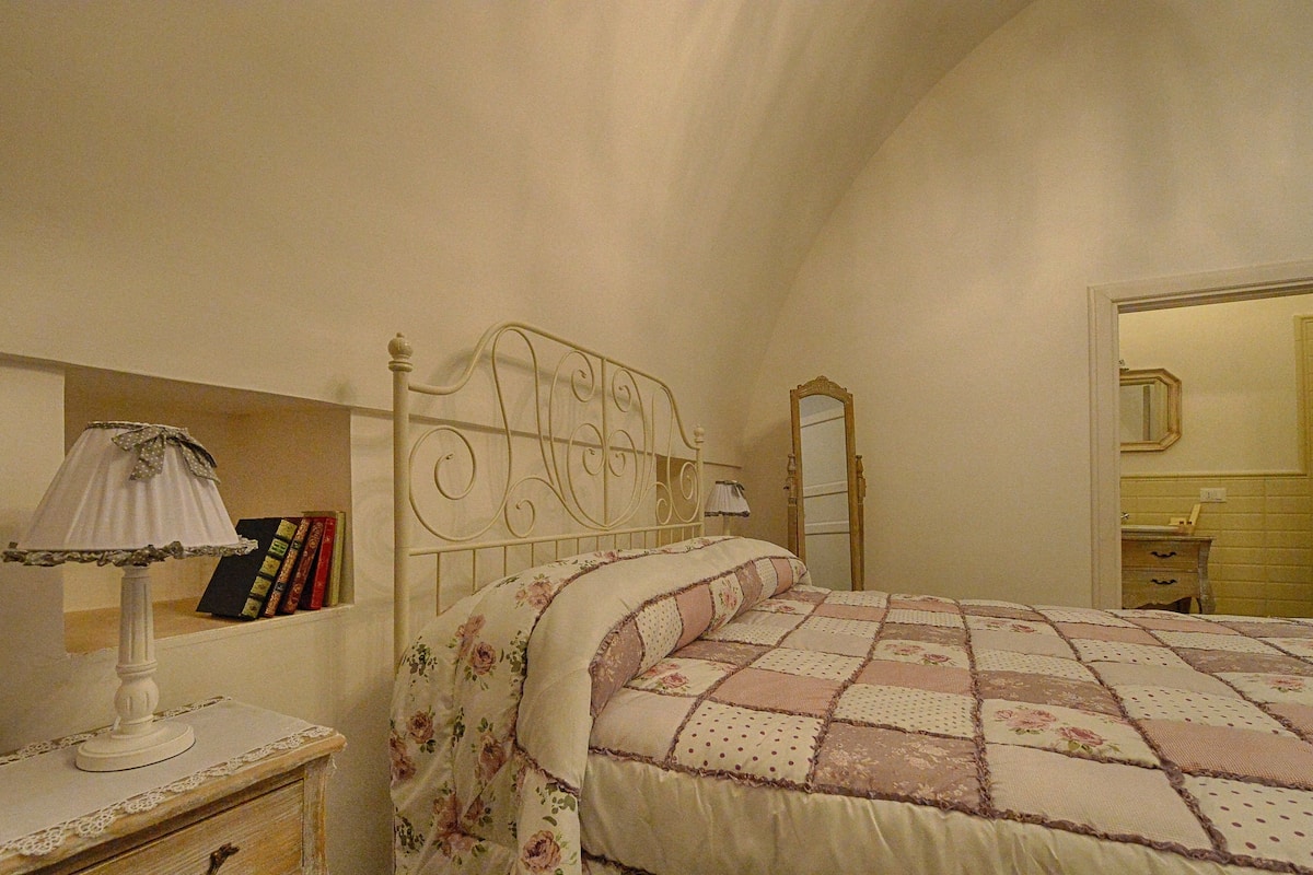 Une chambre à coucher avec un lit, une table de chevet avec une lampe, un miroir et une étagère avec des livres.