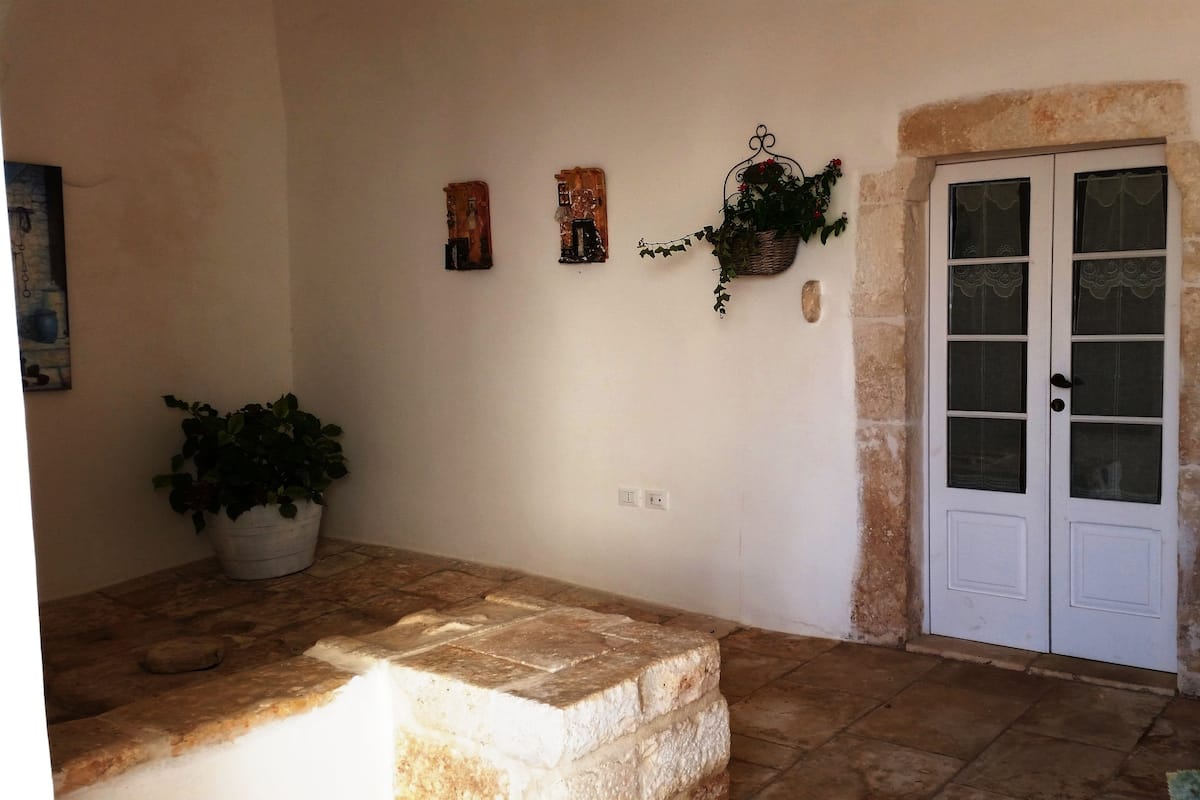 Un couloir au sol dallé de pierre, avec une porte blanche, des plantes en pot et des décorations murales.