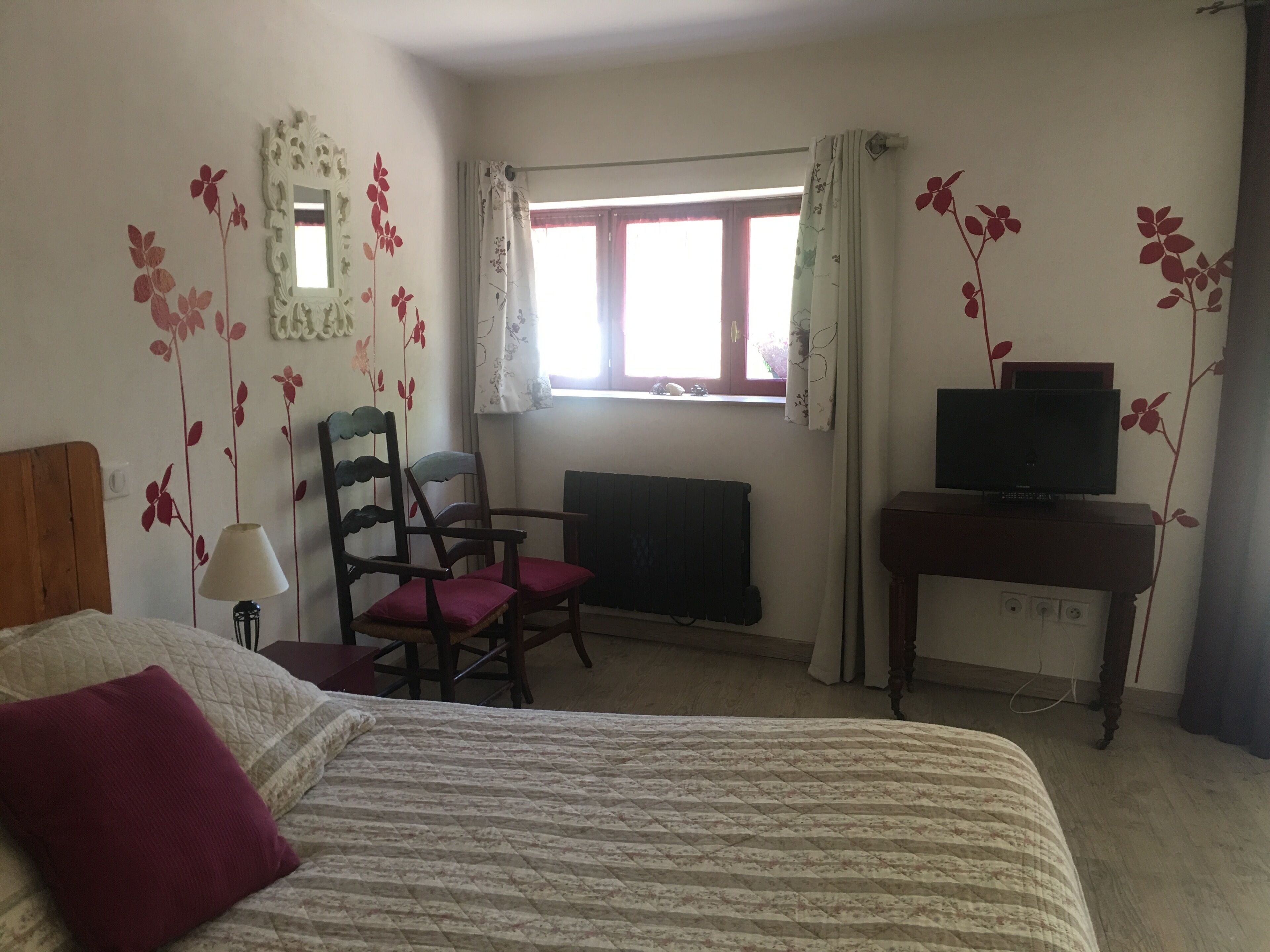 Une chambre à coucher comprenant un lit, deux chaises, un bureau avec une télévision, une fenêtre avec des rideaux et des éléments de décoration murale.