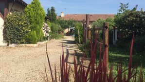 Un camino de grava conduce a una casa rústica con techo de tejas, rodeada de exuberante vegetación y un portón de madera.