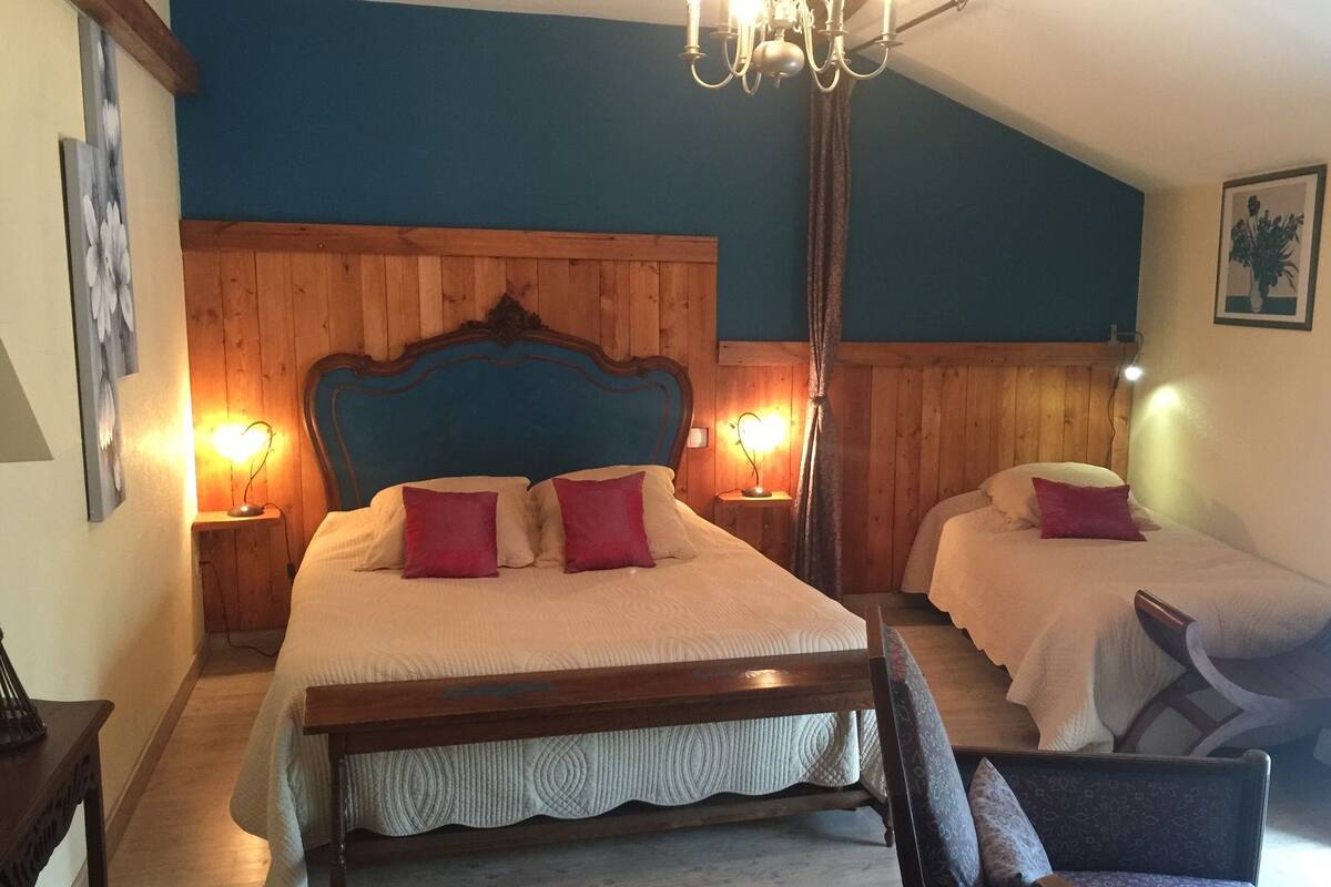 Un dormitorio con cabecera de madera, dos camas con ropa de cama blanca y almohadas rojas, una araña y una mesita de noche con lámpara.