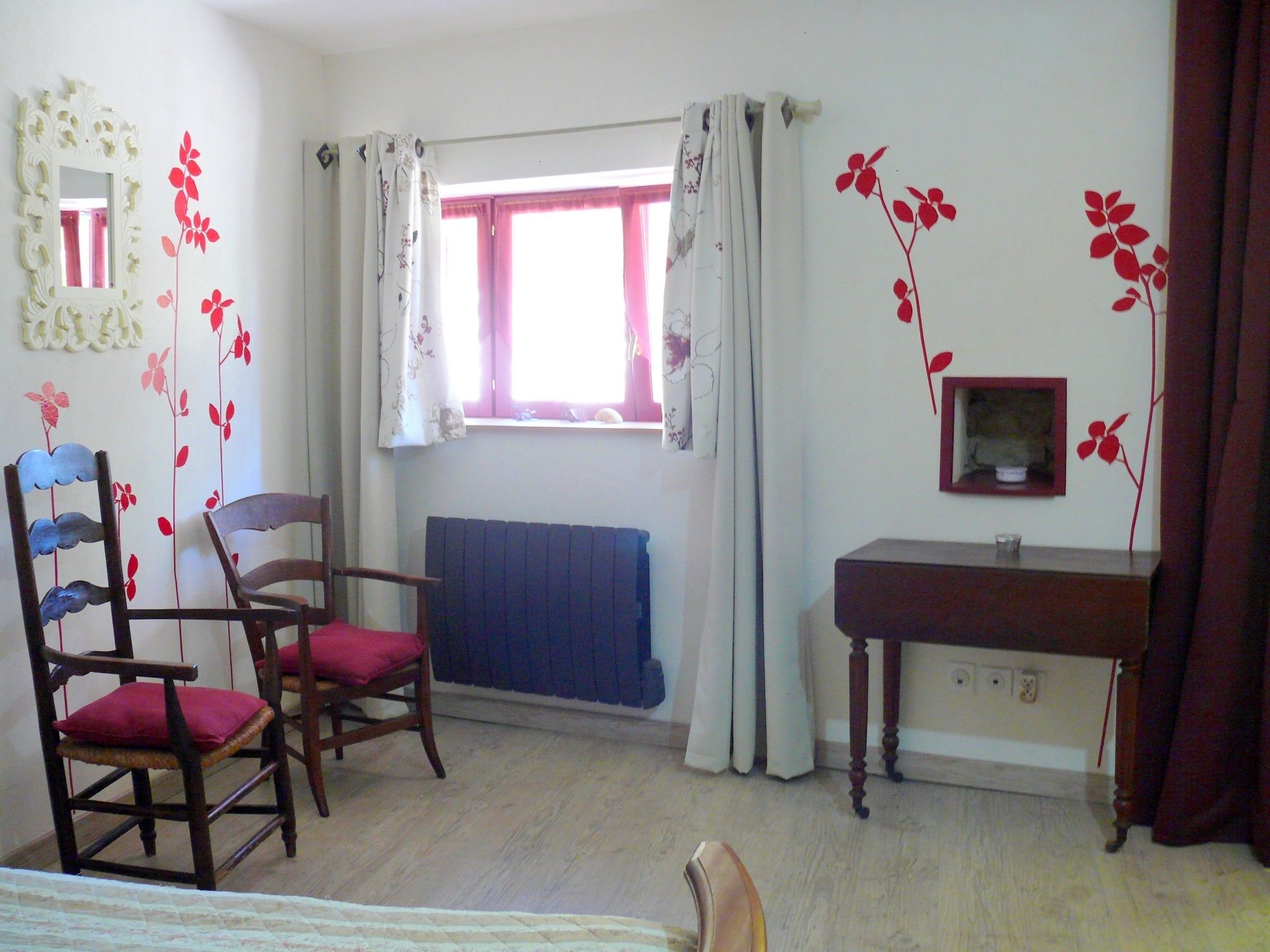 A room with two wooden chairs with red cushions, a small table, a mirror, and a window with curtains.