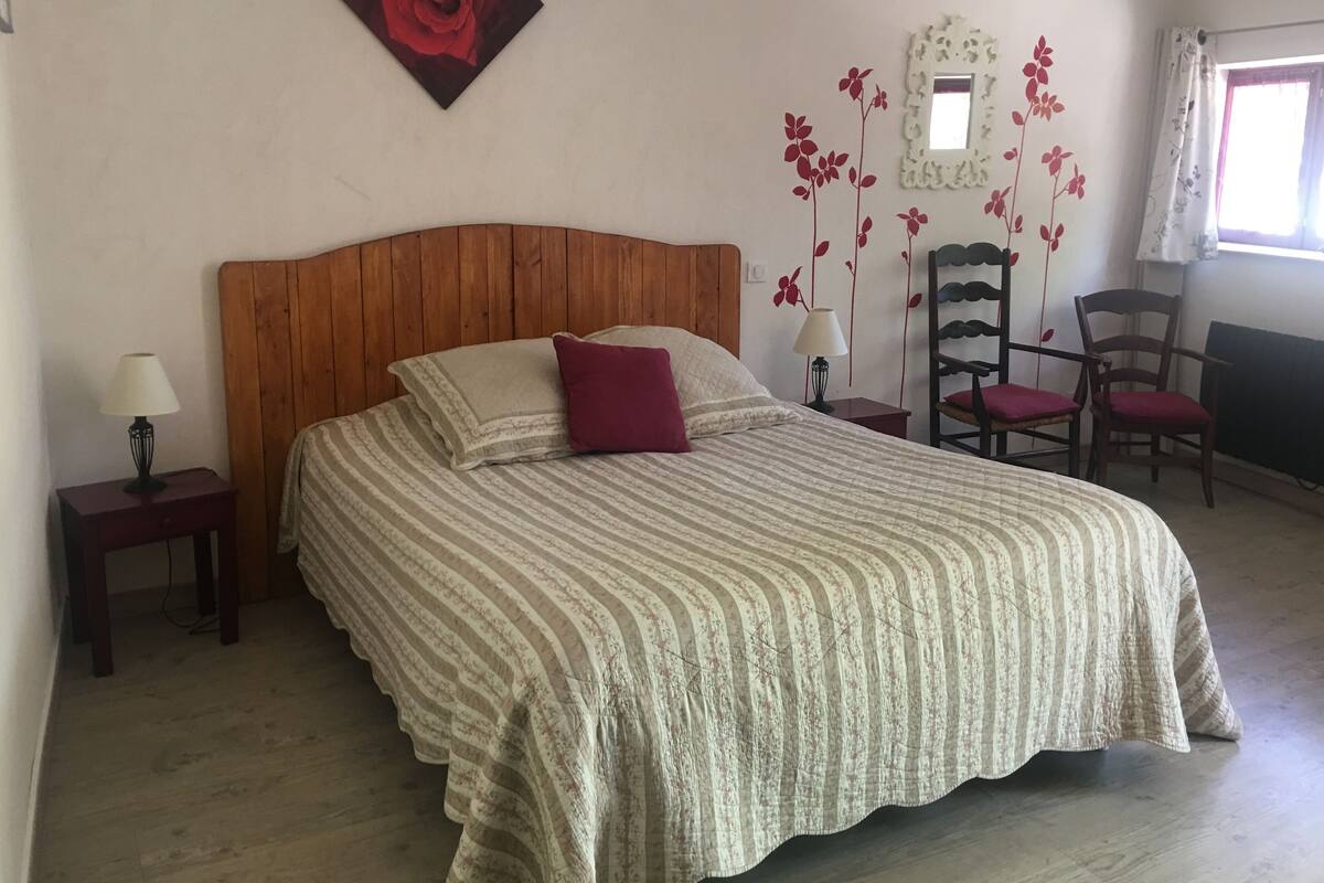 Un dormitorio con cabecera de madera, cama con ropa de cama a rayas, una decoración de rosa roja, un espejo y dos sillas.