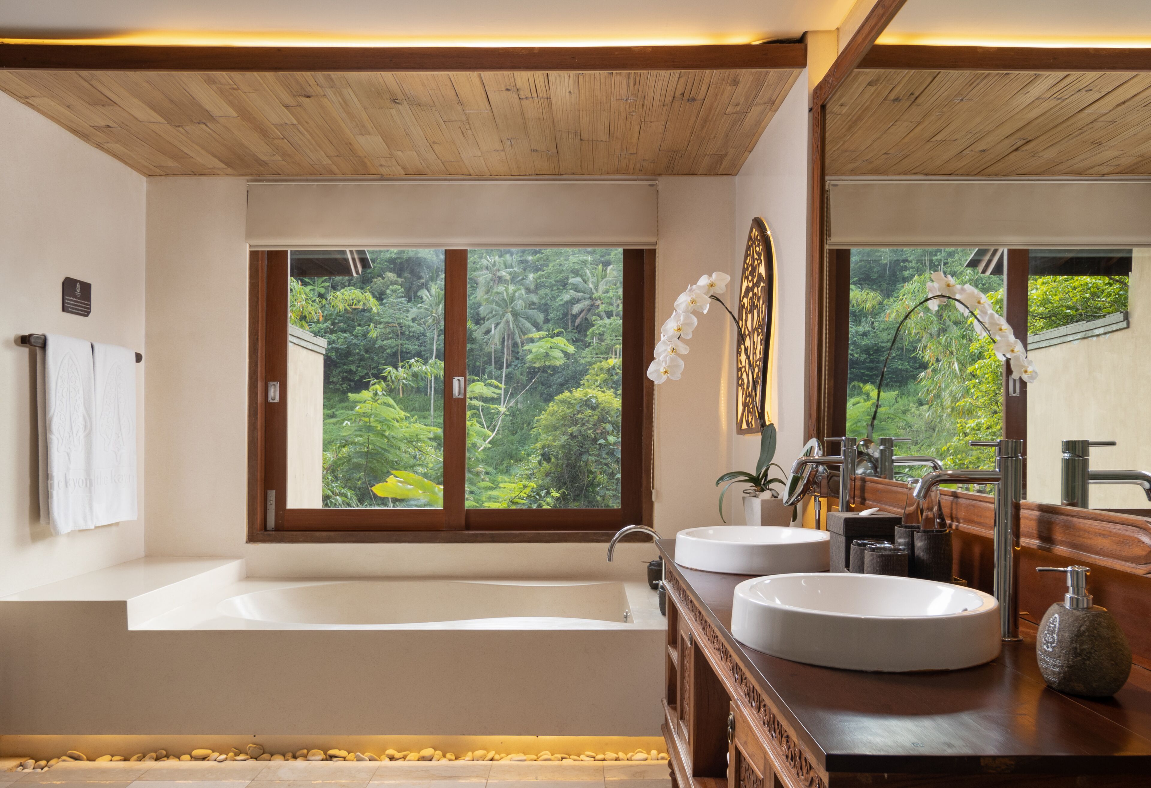 A modern bathroom with a large bathtub, two sinks, and a view of lush greenery outside.
