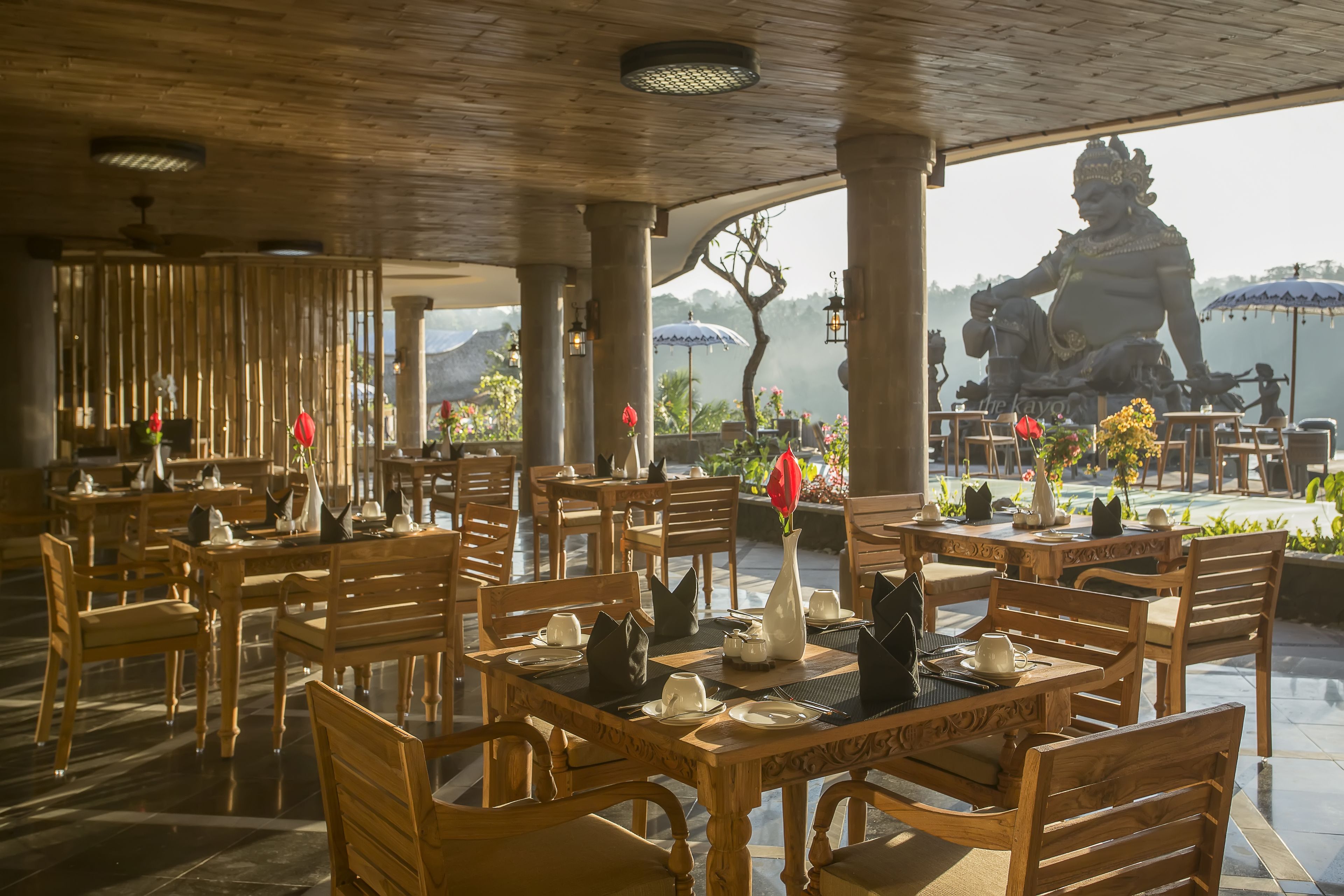 A restaurant with wooden furniture, tables set for dining, and a statue in the background.