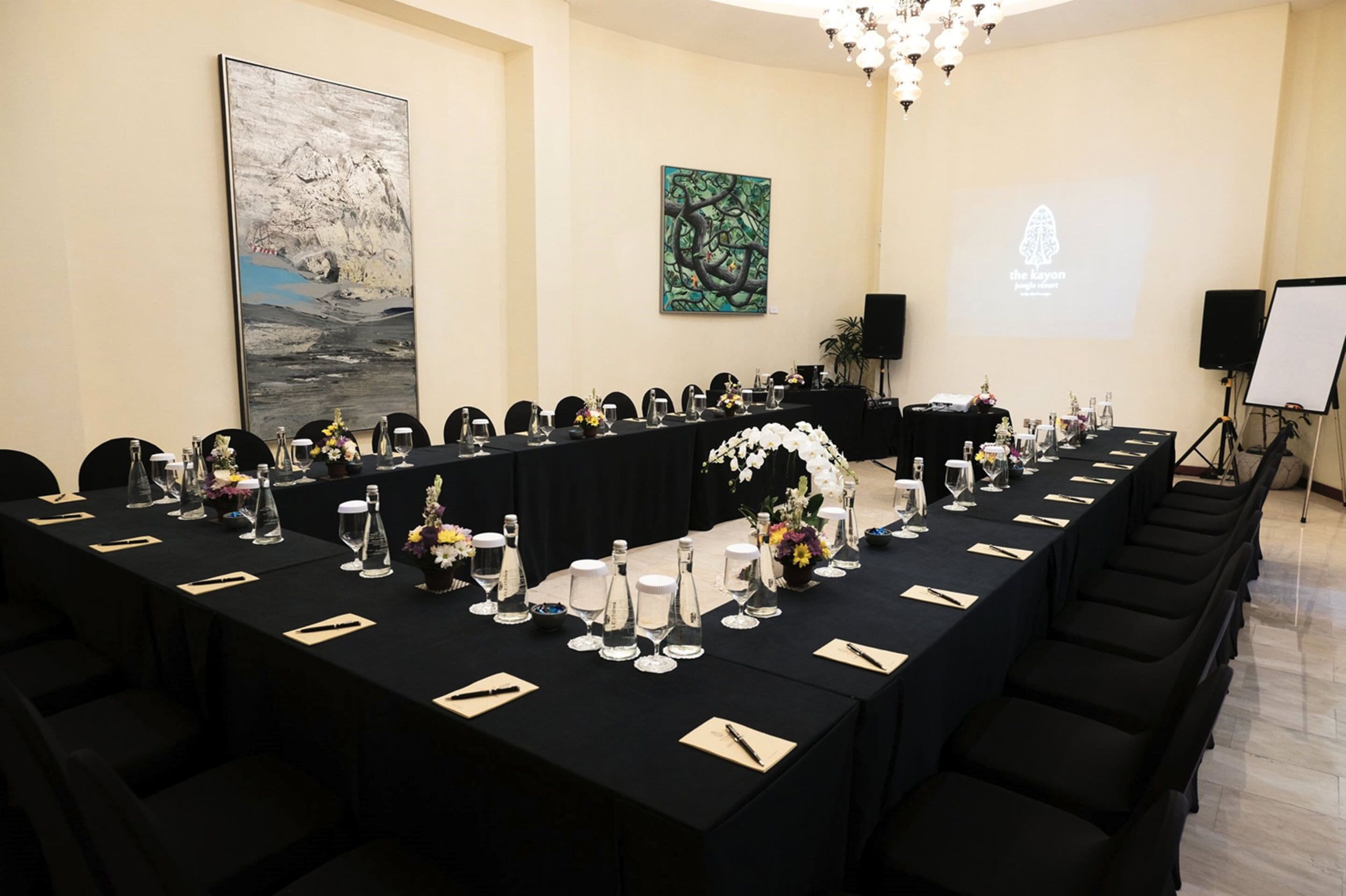 Un salón de conferencias con una larga mesa dispuesta para una reunión, decorada con flores y vasos de agua.