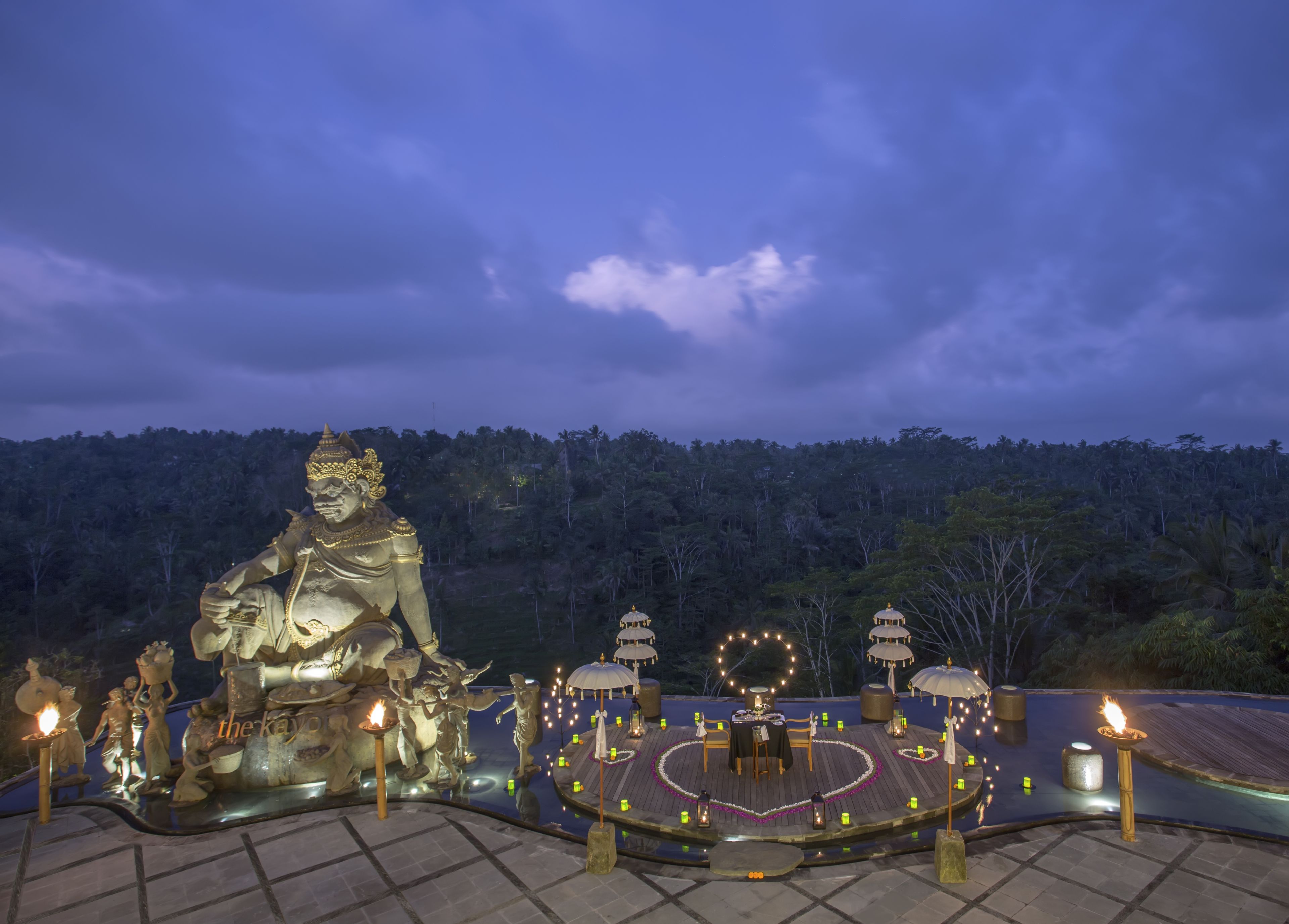 A large Buddha statue surrounded by a circular arrangement of tables and lanterns, set against a backdrop of a forested hill.