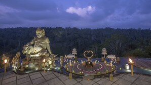 A large Buddha statue surrounded by a circular arrangement of tables and lanterns, set against a backdrop of a forested hill.