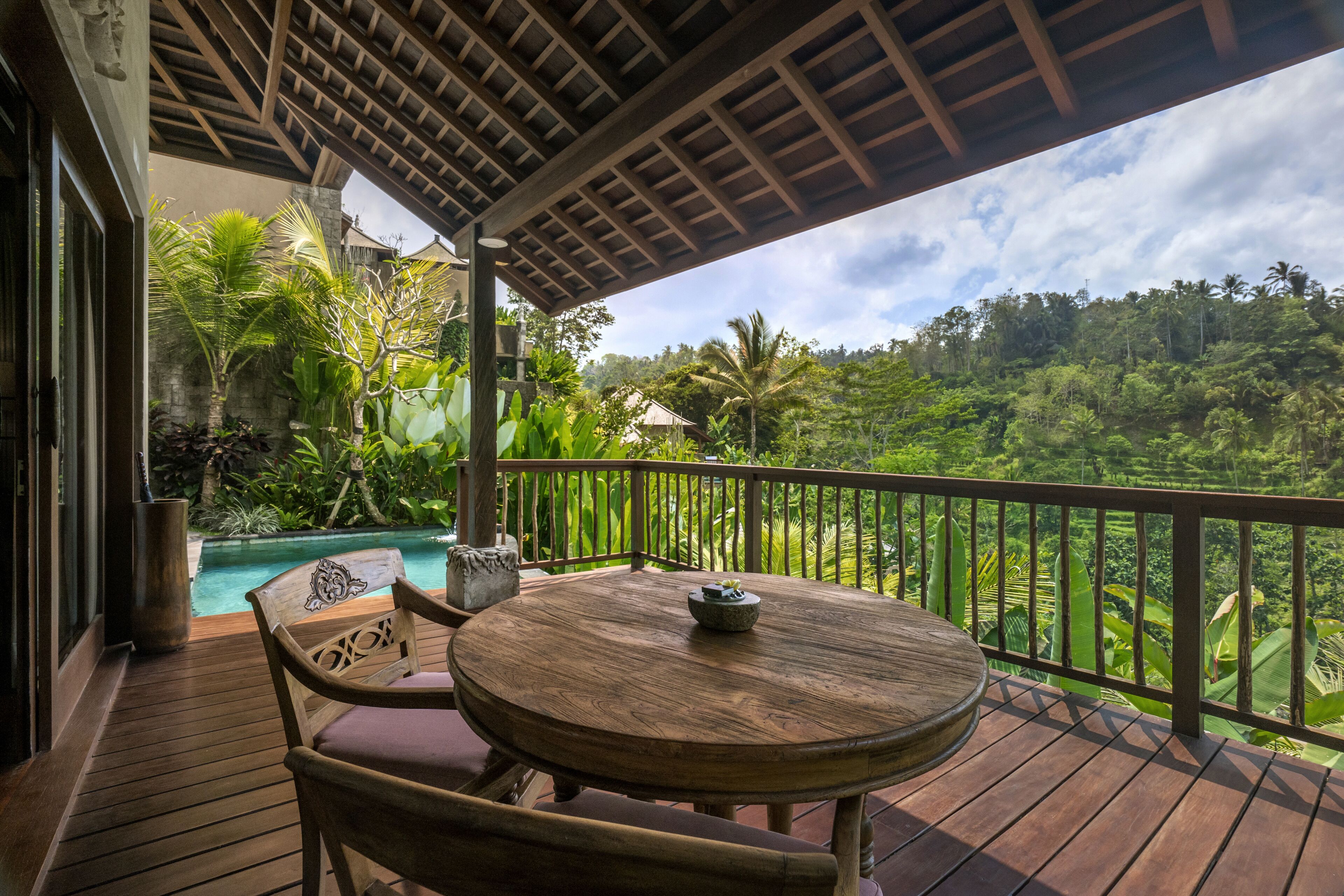 A wooden deck with a round table and chairs, overlooking a lush garden and a pool.