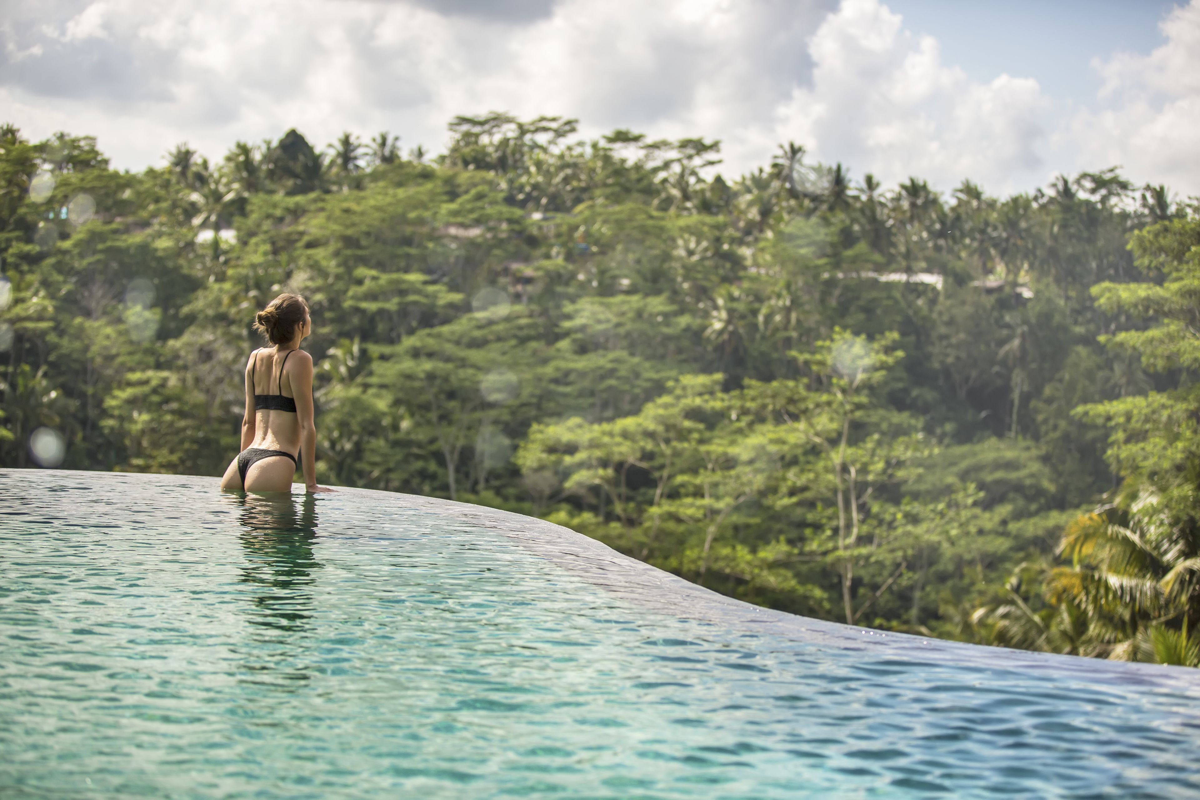 A woman sitting on the edge of a clear pool surrounded by lush greenery.