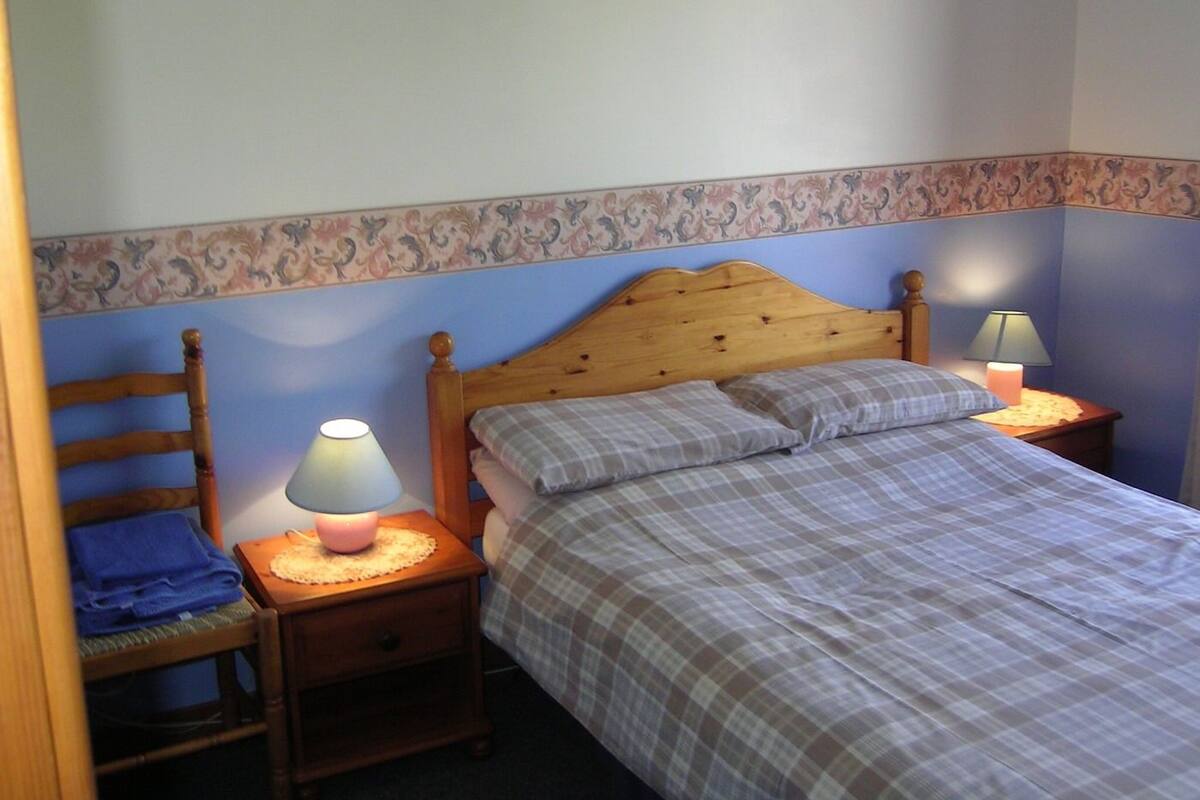 Une chambre à coucher avec un lit en bois, deux tables de chevet, une chaise et un mur décoré d’un motif.