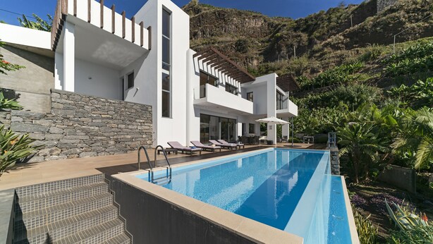 A modern poolside area with a clear blue pool, steps leading to a white building, and a stone wall. - The Designhouse by Our Madeira (Calheta)