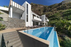 A modern poolside area with a clear blue pool, steps leading to a white building, and a stone wall. - The Designhouse by Our Madeira (Calheta)