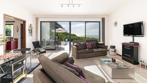 A modern living room with a sofa, coffee table, and TV. There is a glass door leading to a balcony with a view of greenery and a mountain. - The Designhouse by Our Madeira (Calheta)