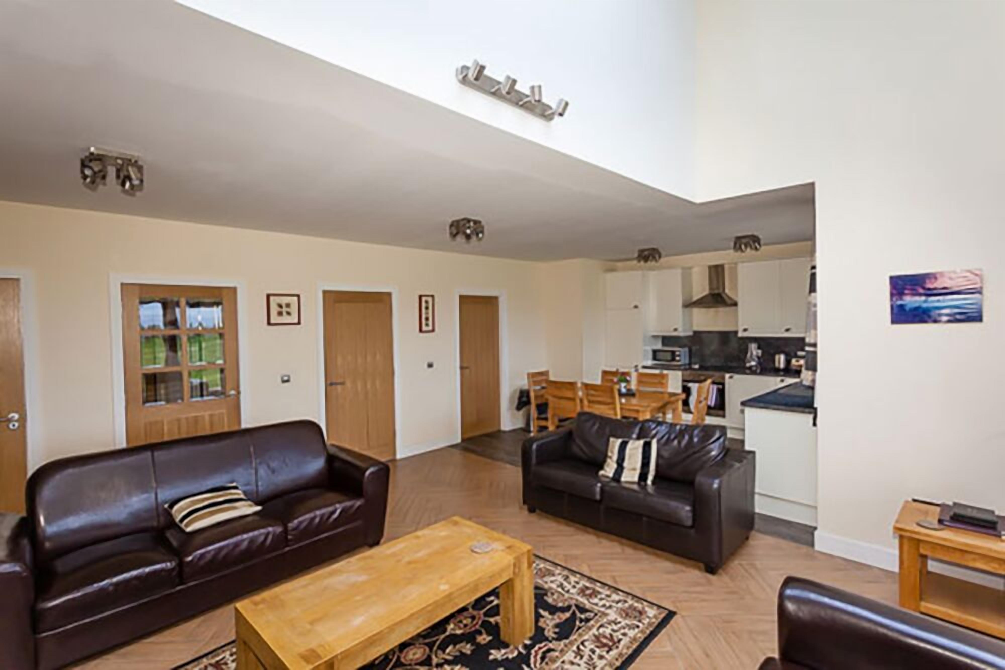 A spacious living room with a brown leather sofa, a wooden coffee table, and a patterned rug. There is a kitchen area visible in the background with white cabinets and a black countertop.