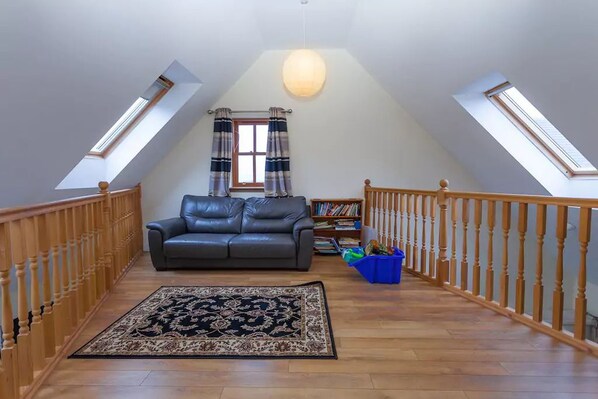 Une chambre à coucher aménagée dans un espace mansardé, avec un plafond en pente, un canapé en cuir noir, un tapis à motifs et une rambarde en bois.