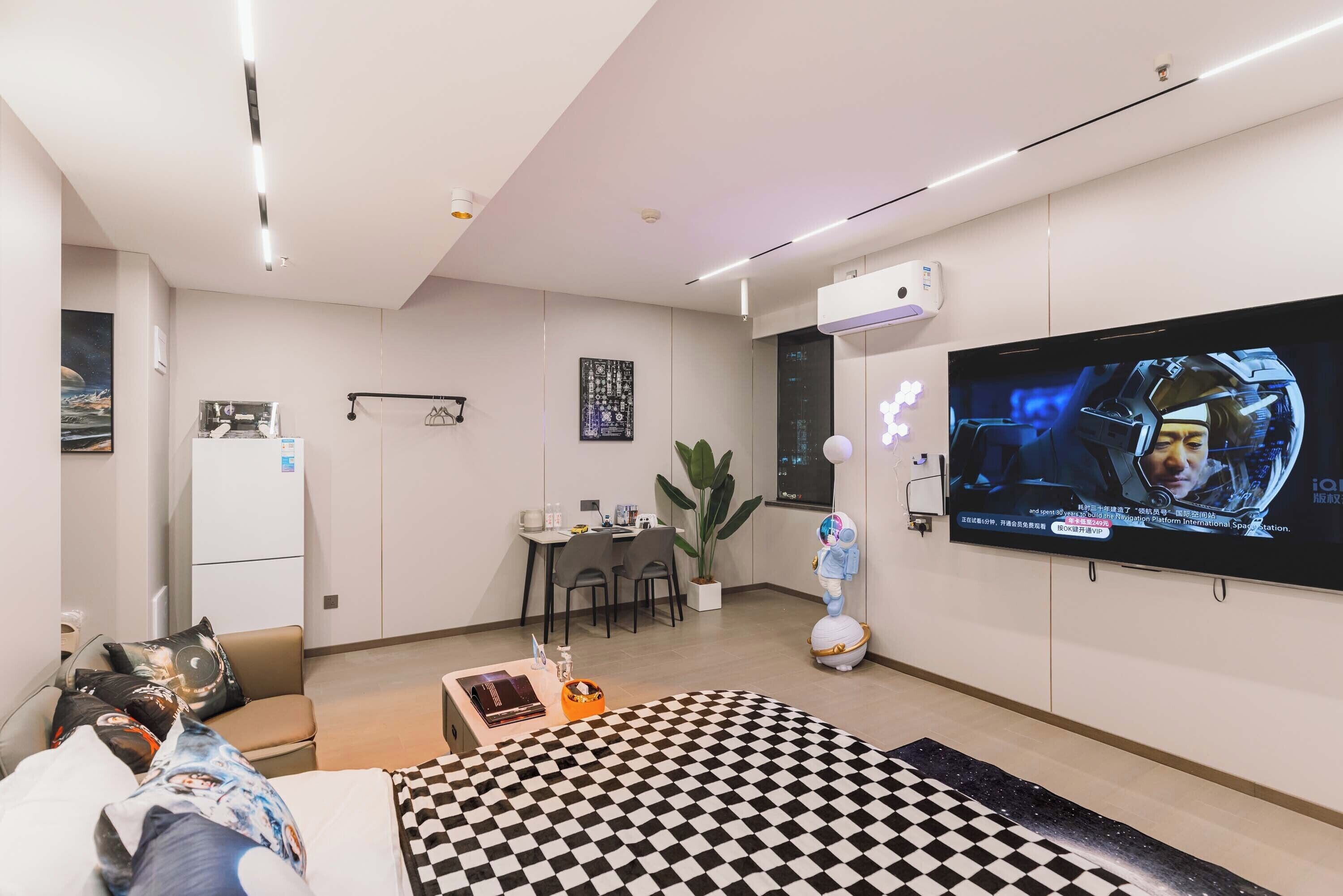 Una habitación de hotel moderna con un televisor de pantalla plana grande, una cama con manta a cuadros, un escritorio con silla, una mesita con una planta y un equipo de aire acondicionado instalado en la pared.