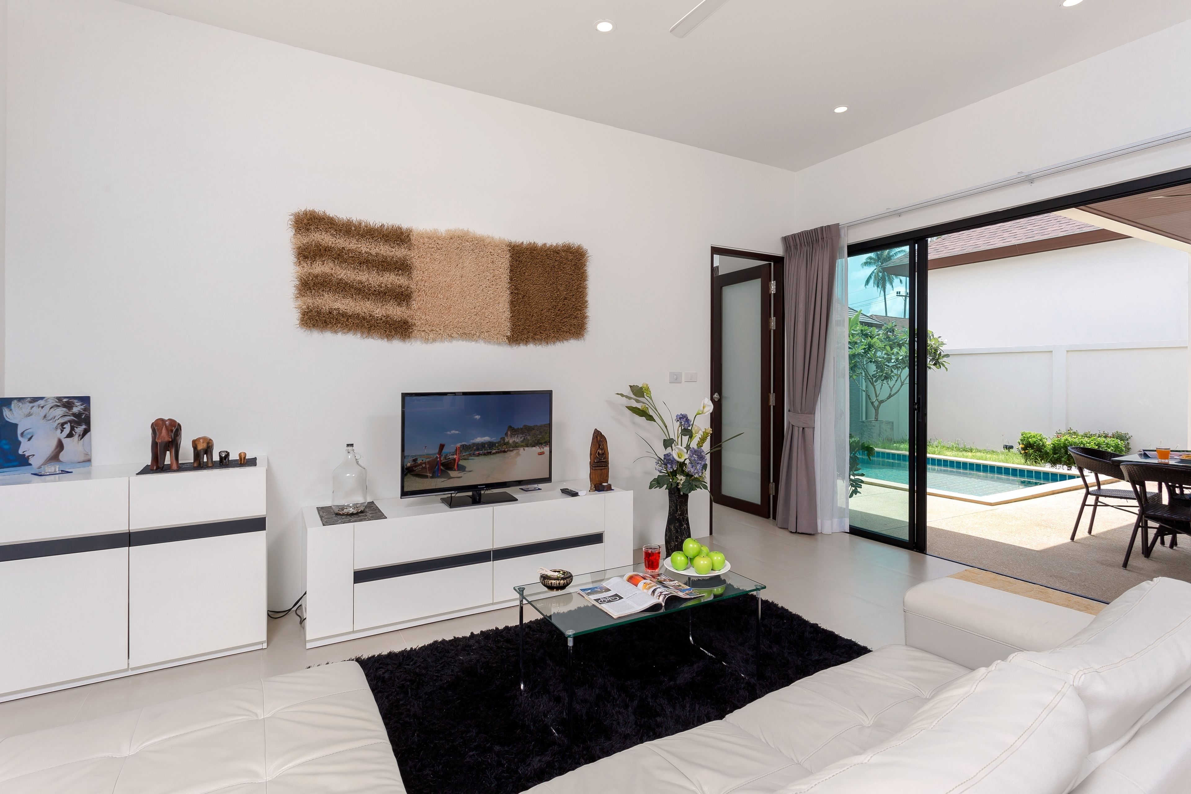 A modern living room with a flat-screen TV, white furniture, a glass coffee table, and a view of an outdoor swimming pool.