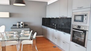 Een moderne keuken met een glazen eettafel, witte stoelen, grijze kasten en roestvrijstalen apparatuur.