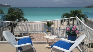 Un balcone con due poltrone in vimini e un tavolino, con vista su una spiaggia e acque turchesi cristalline, con una nave all'orizzonte.