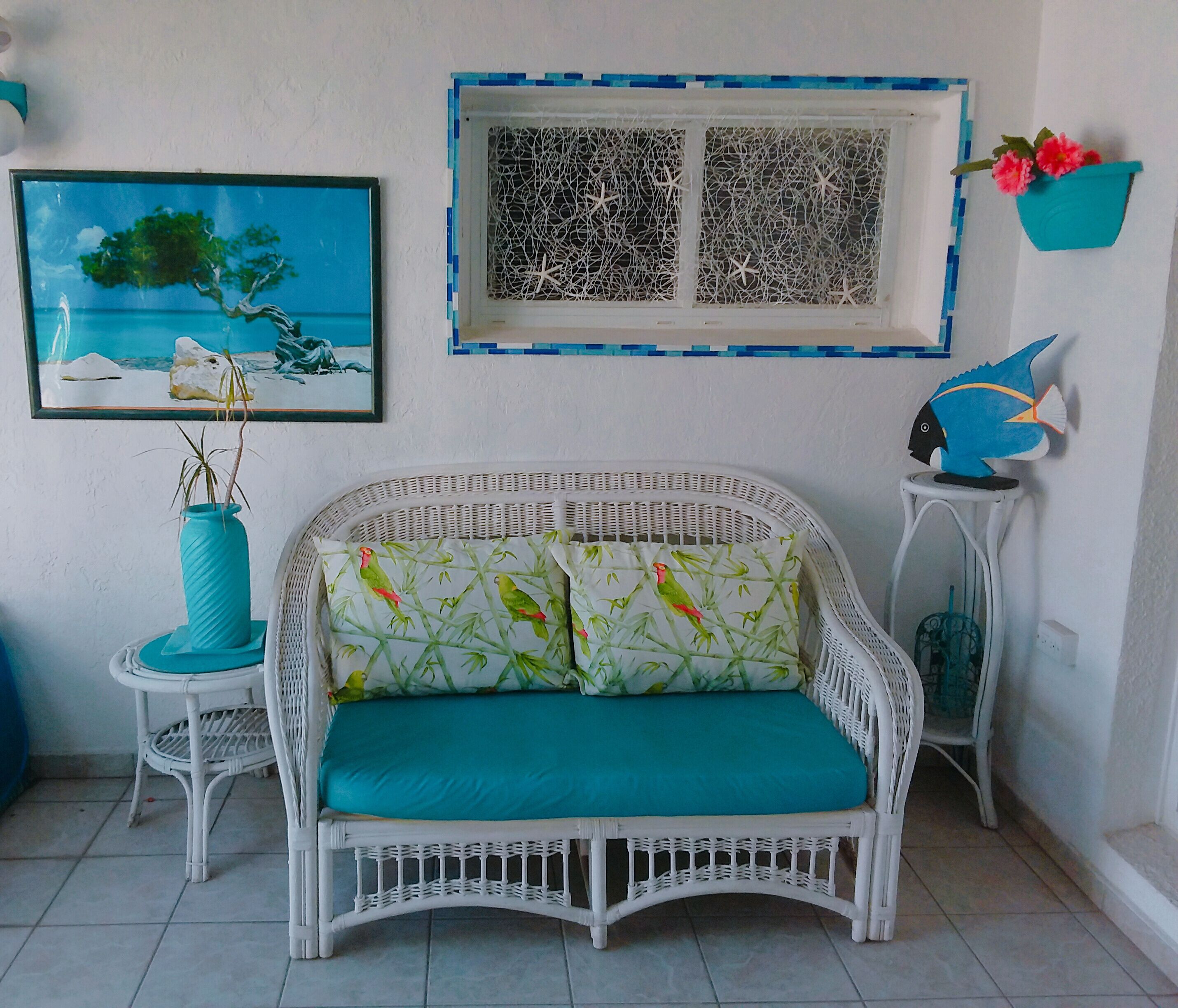 Un canapé en osier blanc agrémenté de coussins turquoise, un tableau encadré représentant un arbre, une fenêtre à cadre bleu et une petite décoration en forme de poisson bleu posée sur un petit meuble blanc.