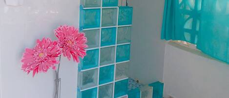 A bathroom with a shower, a hand sanitizer dispenser, a blue mat, and pink flowers.
