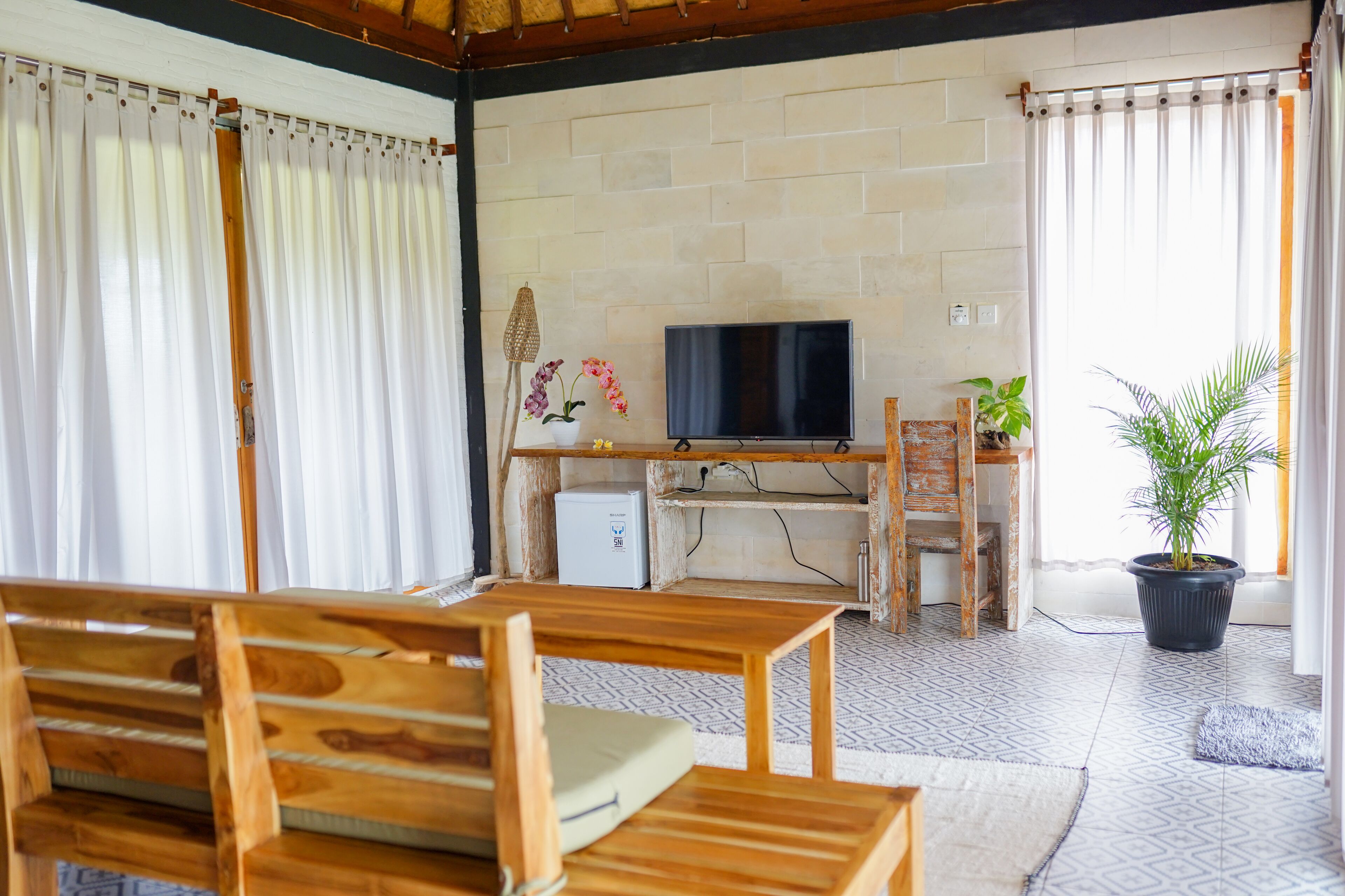 A cozy living area with a wooden sofa, a TV on a stand, a small refrigerator, and a potted plant.