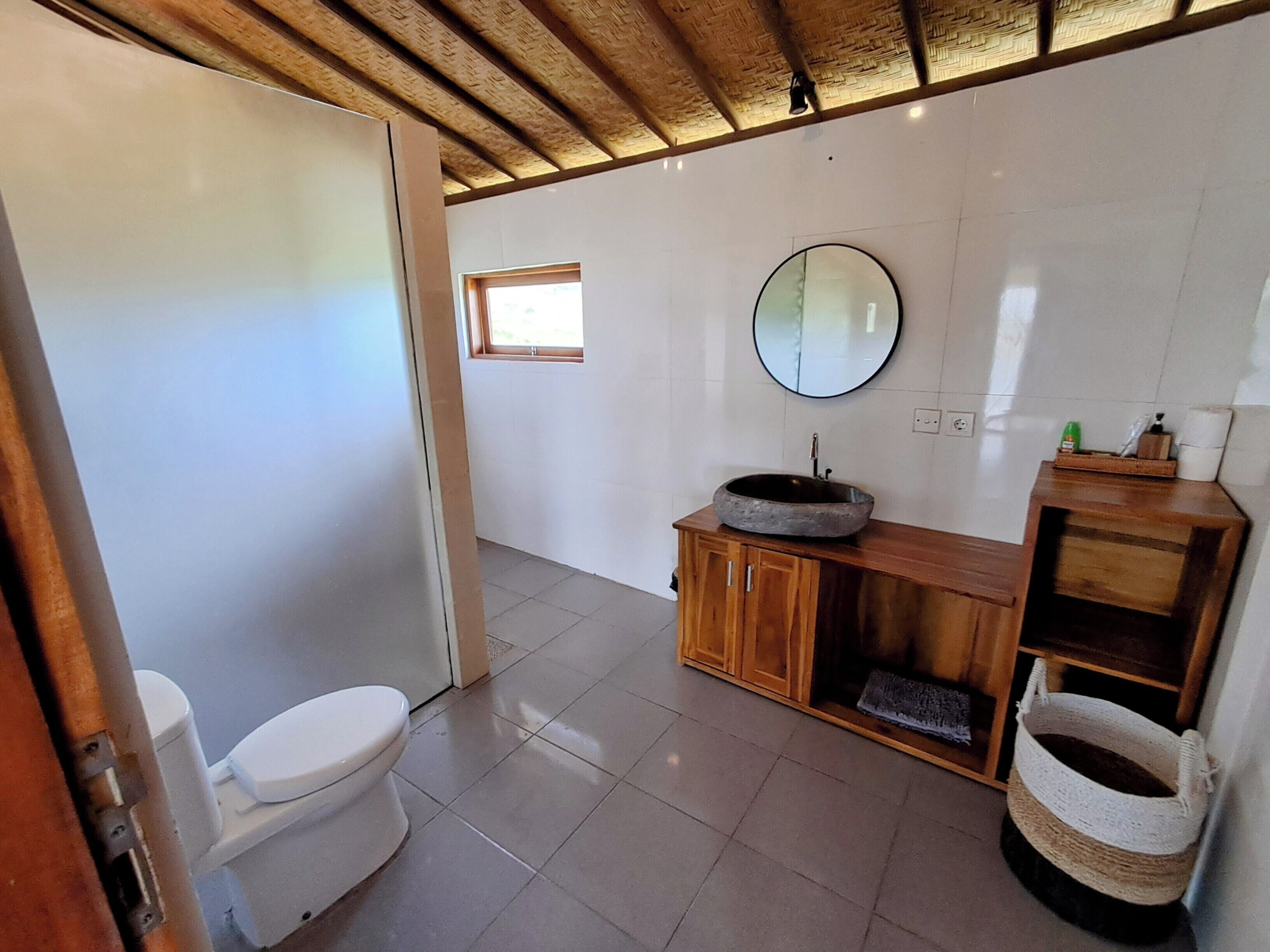 A bathroom with a toilet, a wooden vanity with a round sink, a mirror, and a small window.