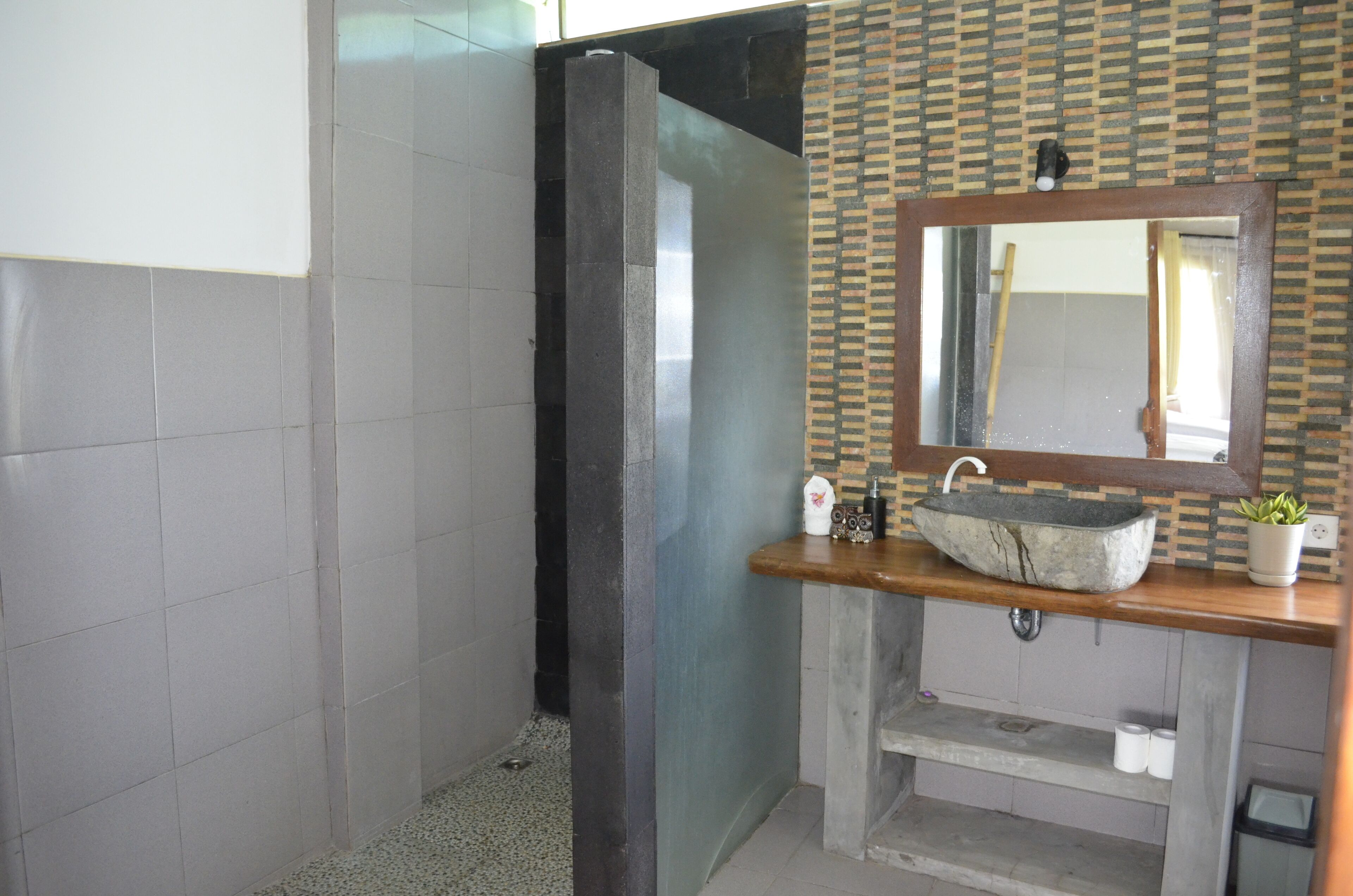 A bathroom with a tiled floor, a shower area, a sink with a stone basin, a mirror, and a small shelf with toiletries.