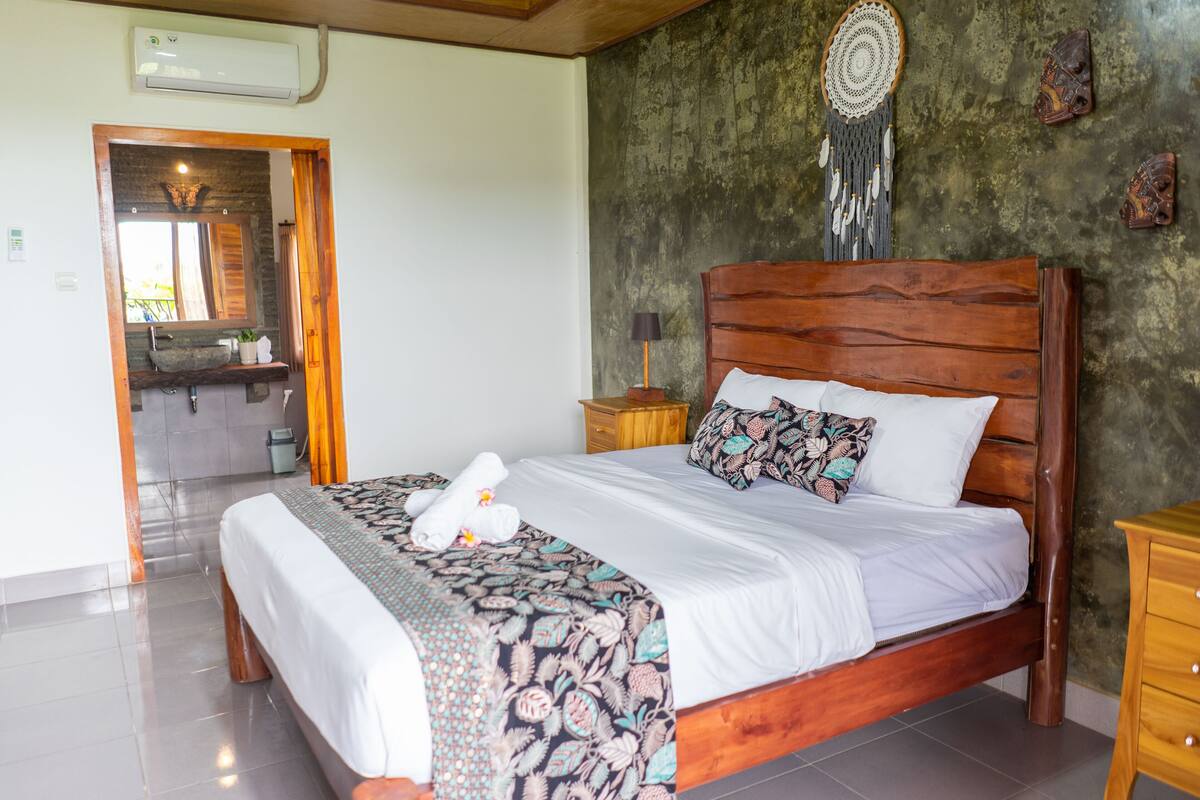 Ein Schlafzimmer mit einem hölzernen Bett, einem Nachttisch, einem an der Wand befestigten Traumfänger und einem durch eine offene Tür einsehbaren Badezimmer.