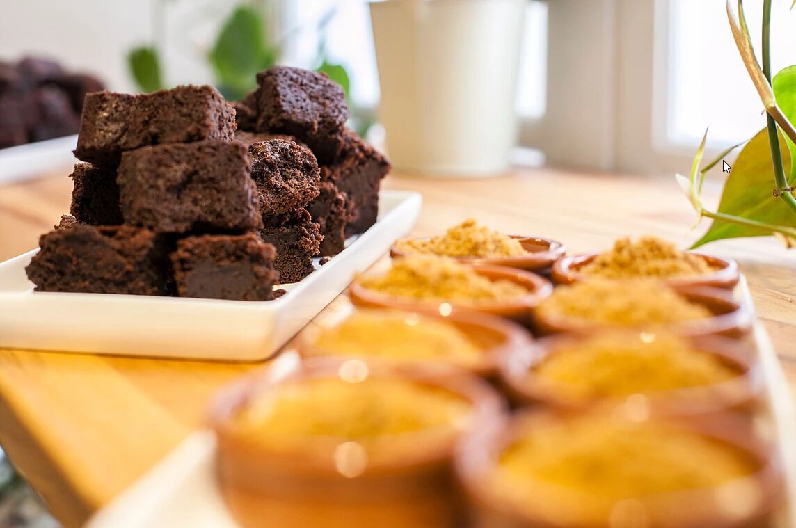 Una bandeja de brownies y varios recipientes de azúcar glas sobre una mesa.