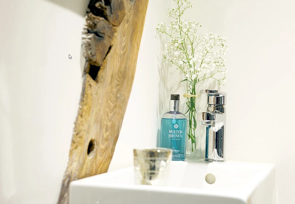 Une salle de bain avec un lavabo blanc, deux bouteilles de shampoing Molton Brown London, un vase transparent contenant des fleurs blanches et un mur à l’aspect bois.