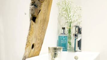 Une salle de bain avec un lavabo blanc, deux bouteilles de shampoing Molton Brown London, un vase transparent contenant des fleurs blanches et un mur à l’aspect bois.