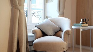 Un sillón color beige con un cojín, situado junto a una ventana con cortinas, una pequeña mesa con un libro y una jarrón metálico, y un armario de madera al fondo.
