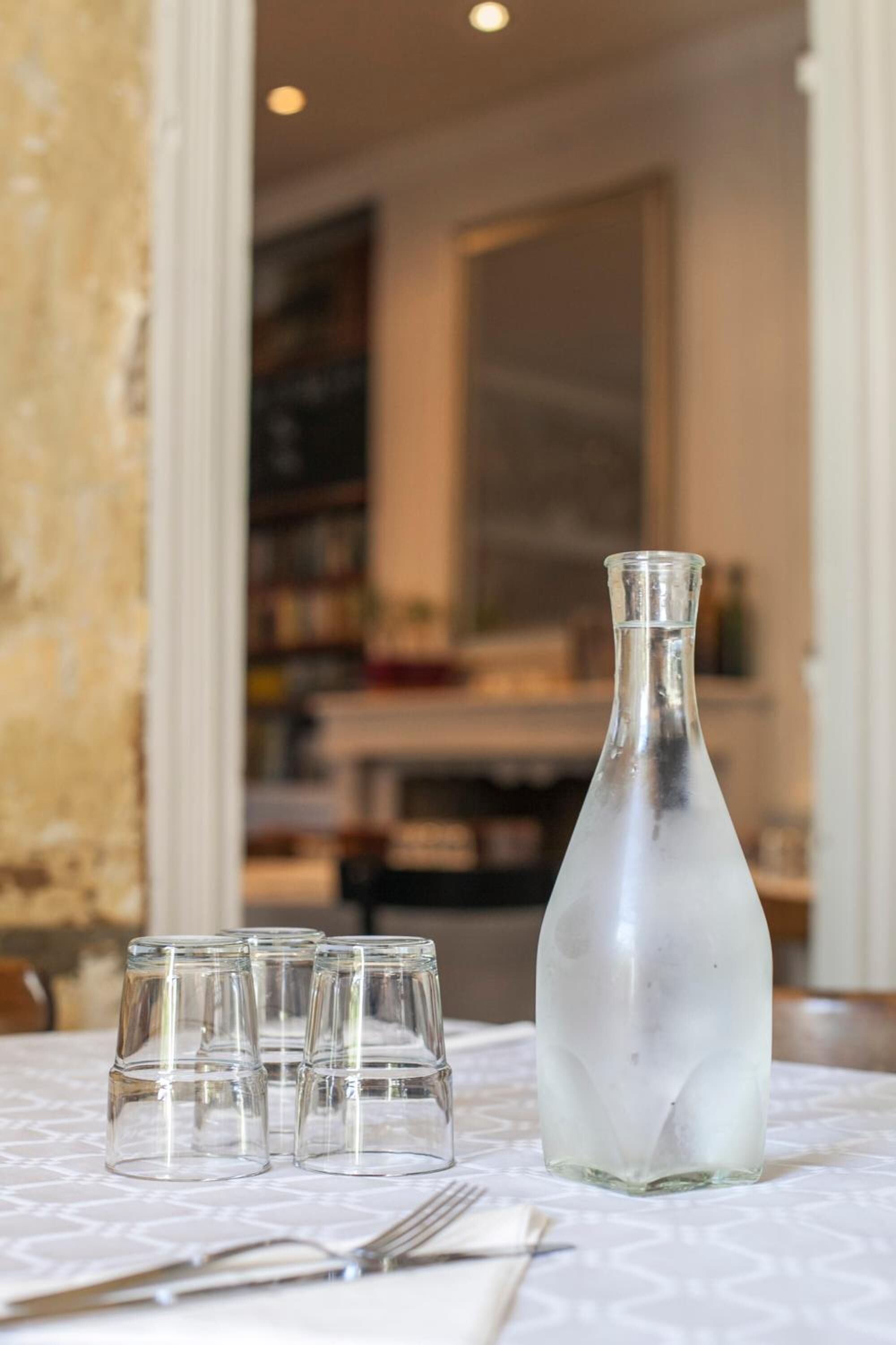 A table with a clear glass bottle and four small glasses, likely for water or a beverage, in a room with a patterned tablecloth and a visible bookshelf in the background.