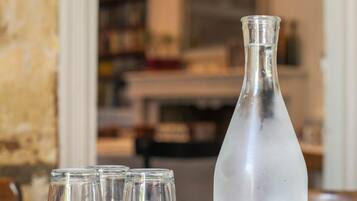 A table with a clear glass bottle and four small glasses, likely for water or a beverage, in a room with a patterned tablecloth and a visible bookshelf in the background.