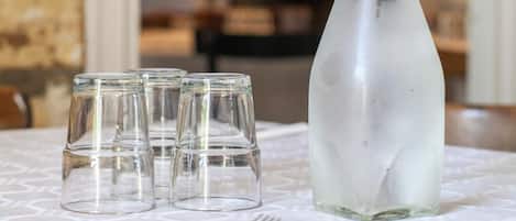 Una mesa con una botella de vidrio transparente y cuatro vasos pequeños, probablemente para agua u otra bebida, en una habitación con un mantel estampado y un estante de libros a la vista.