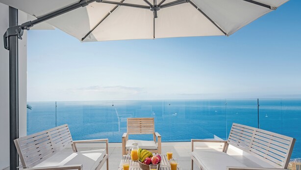 Een balkon met een tafeltje voor twee personen, compleet met een fruitmand en glazen sap, met uitzicht op een heldere blauwe zee met boten.