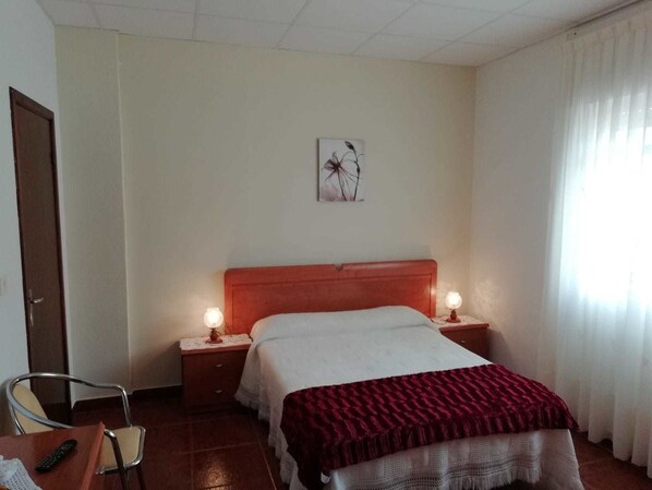 A hotel room with a bed, two bedside lamps, a chair, a desk with a remote control, and a door. - Pension Porto Carral (Muros)