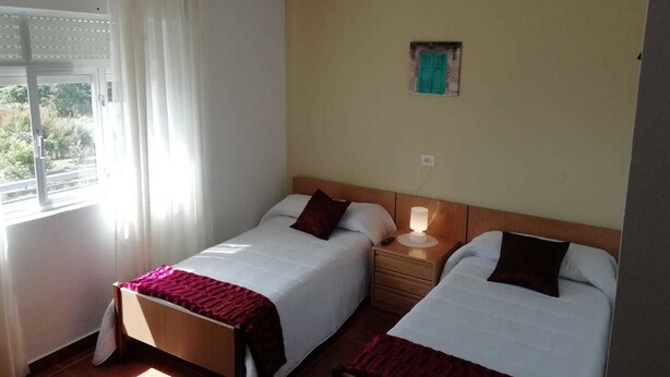 A hotel room with two single beds, a nightstand with a lamp, a window with curtains, and a small wall decoration. - Pension Porto Carral (Muros)