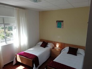 A hotel room with two single beds, a nightstand with a lamp, a window with curtains, and a small wall decoration. - Pension Porto Carral (Muros)