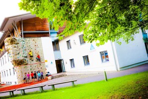 A climbing wall with a group of people climbing, a building with a blue and white facade, and a grassy area with benches. - DJH Garmisch- Partenkirchen - Hostel (Garmisch-Partenkirchen)