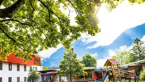 A playground with a slide, picnic tables, and benches in a park with buildings and mountains in the background. - DJH Garmisch- Partenkirchen - Hostel (Garmisch-Partenkirchen)