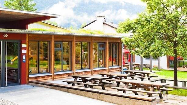 A building with a red exterior, large glass windows, and a covered picnic area with multiple benches. - DJH Garmisch- Partenkirchen - Hostel (Garmisch-Partenkirchen)